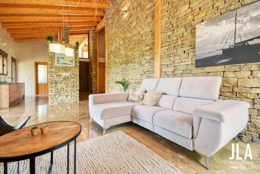 Sala de estar con pared de piedra, sofá claro, alfombra, vigas de madera en el techo y arte.