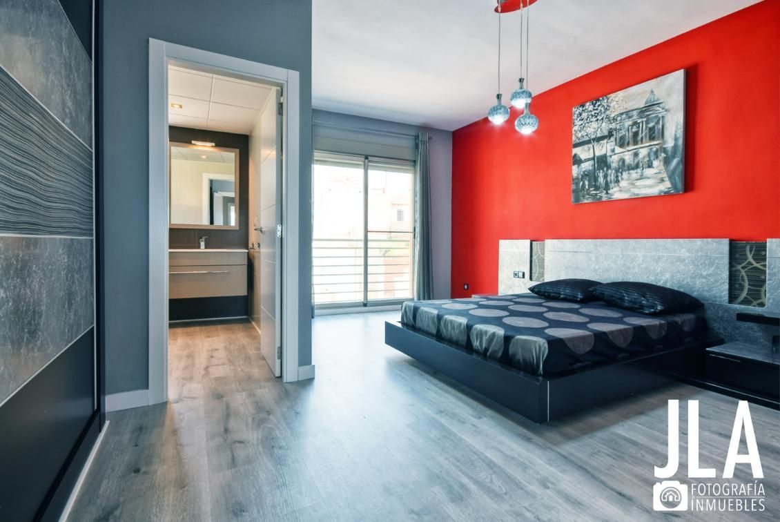 Dormitorio con pared de acento rojo, cama negra y puerta abierta a un baño.