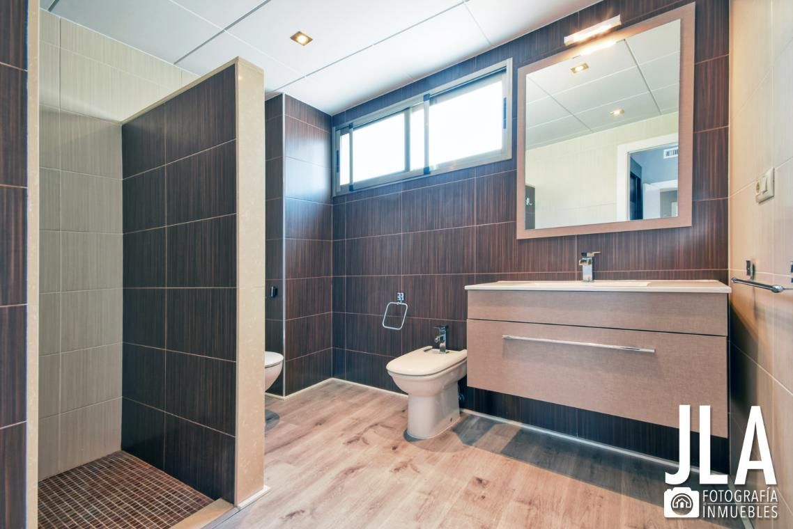 Baño con paredes de azulejos marrones, ducha, lavabo, inodoro y ventana. Suelo imitación madera clara.