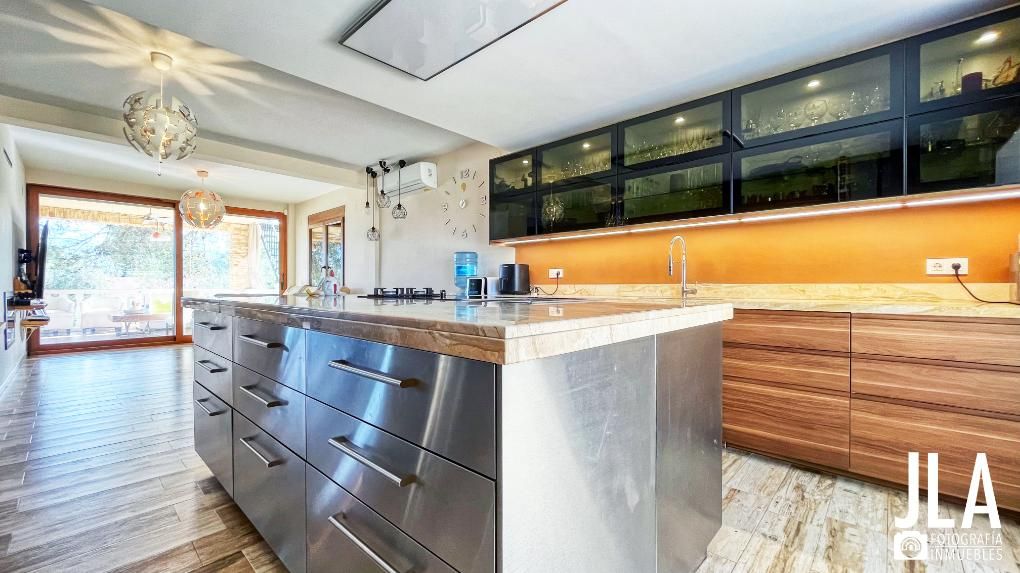 Cocina moderna con isla de acero inoxidable, gabinetes de madera y gabinetes superiores con frente de vidrio.