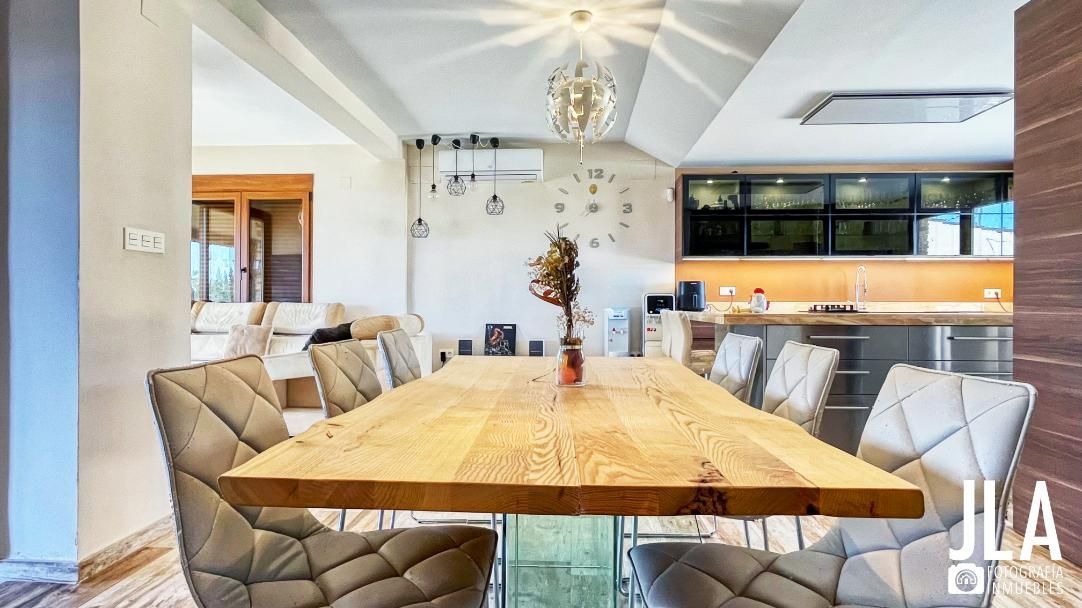 Comedor con mesa de madera, sillas grises y lámpara de araña. Cocina al fondo.