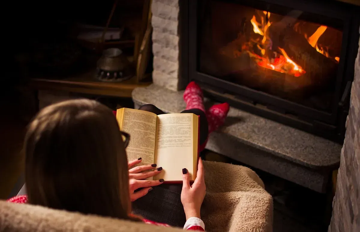 Una mujer está sentada en una silla leyendo un libro frente a una chimenea.
