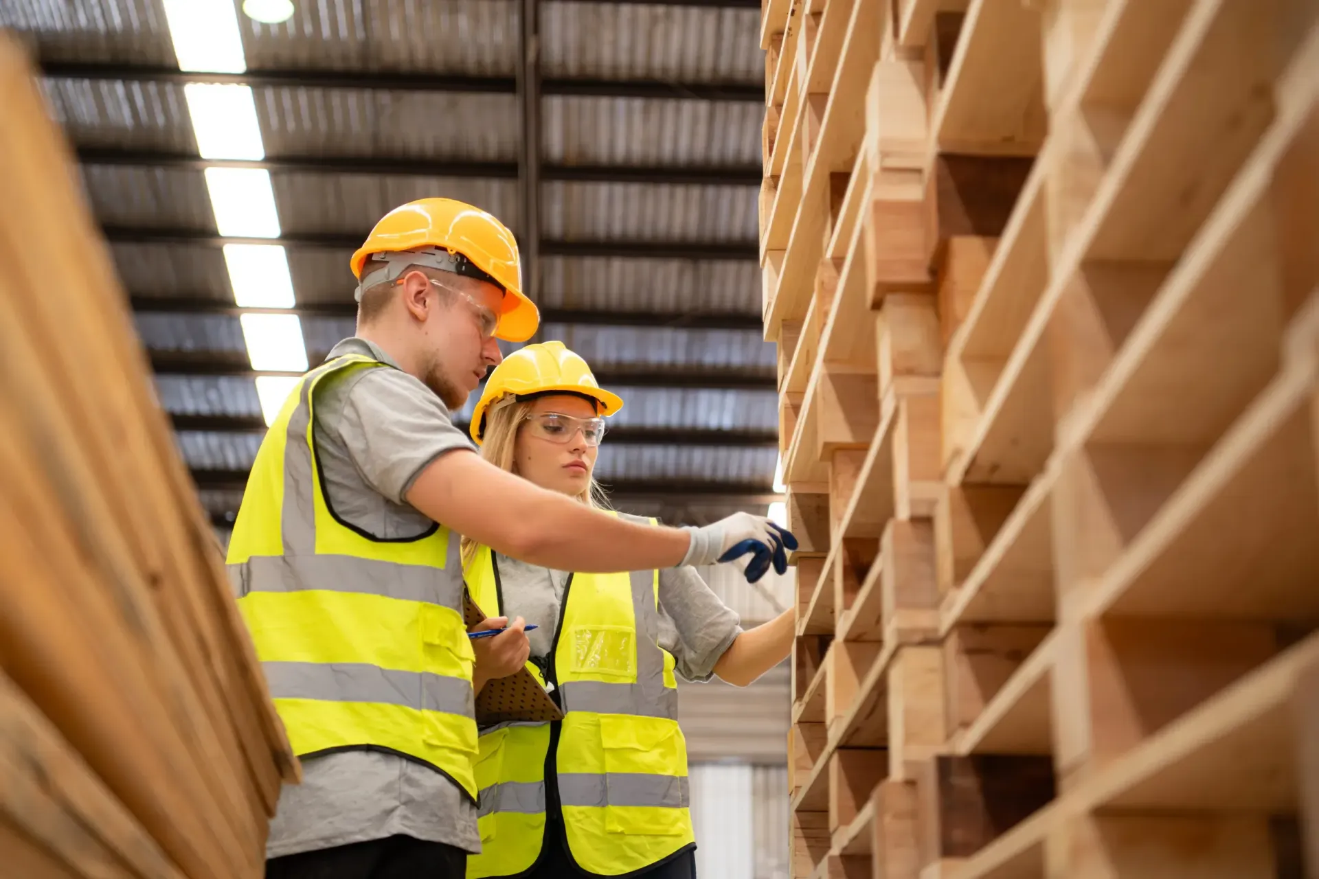 Dos trabajadores con chalecos de seguridad amarillos y cascos inspeccionan una pila de palés de madera en un almacén.