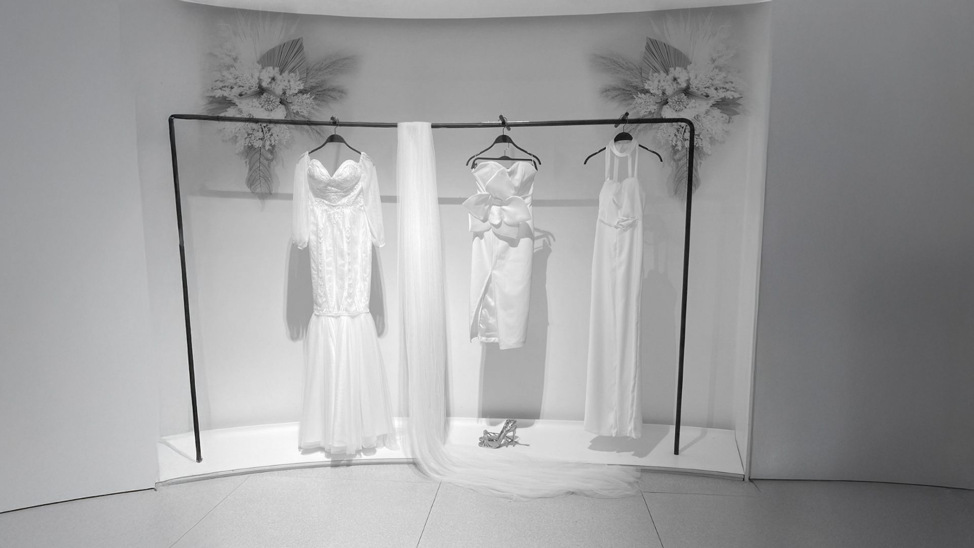 Los vestidos de novia cuelgan en un perchero negro; el fondo incluye arreglos florales y telas drapeadas.