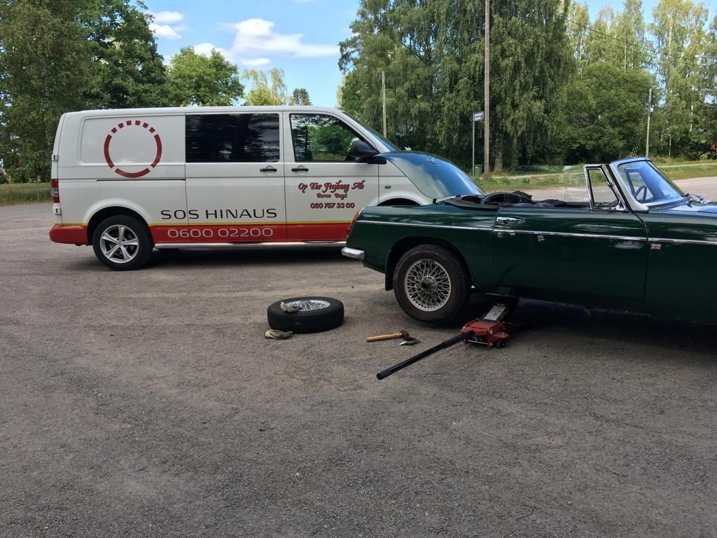 Valkoinen pakettiauto vihreän klassisen avoauton vieressä, jonka konepelti on auki; rengas, tunkki ja työkalut asfaltilla, puita taustalla
