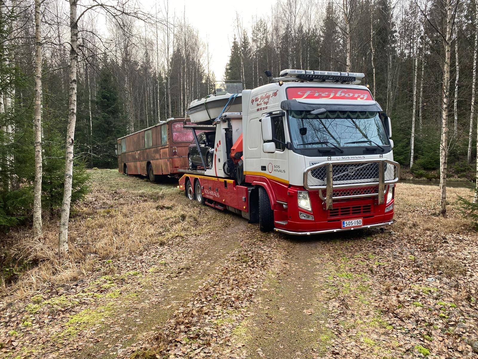 Punainen metsäauto vetää puuperävaunuja hiekkatiellä koivumetsässä