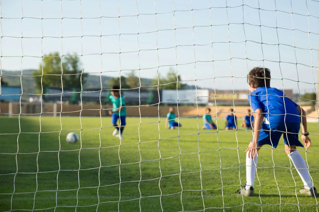 Niño portero con uniforme de fútbol azul, mirando hacia un balón de fútbol en un campo verde, otros niños en el fondo.