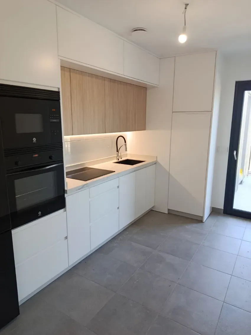 Cocina moderna blanca con horno empotrado negro, suelo de baldosas grises y una ventana sobre el fregadero.