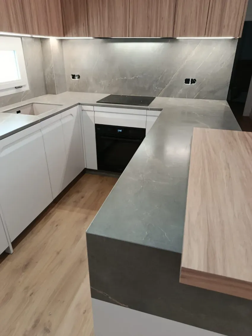 Cocina moderna con gabinetes de madera clara, encimeras blancas, isla gris y electrodomésticos empotrados.