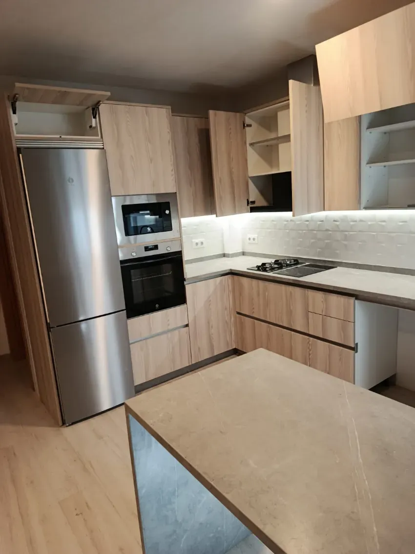 Cocina moderna con nevera de acero inoxidable, horno empotrado, armarios de madera y encimeras beige.
