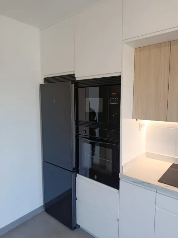 Cocina moderna blanca con nevera alta negra y horno empotrado junto a un mueble de madera clara.