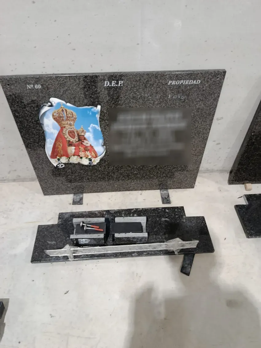 Placa conmemorativa de granito negro con una pegatina navideña y flores en la base sobre un suelo de hormigón.