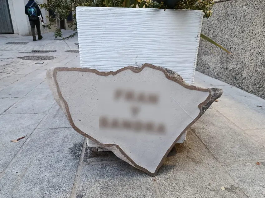 Cartel de piedra rota con texto borroso en una zona pavimentada al aire libre, con una persona al fondo.