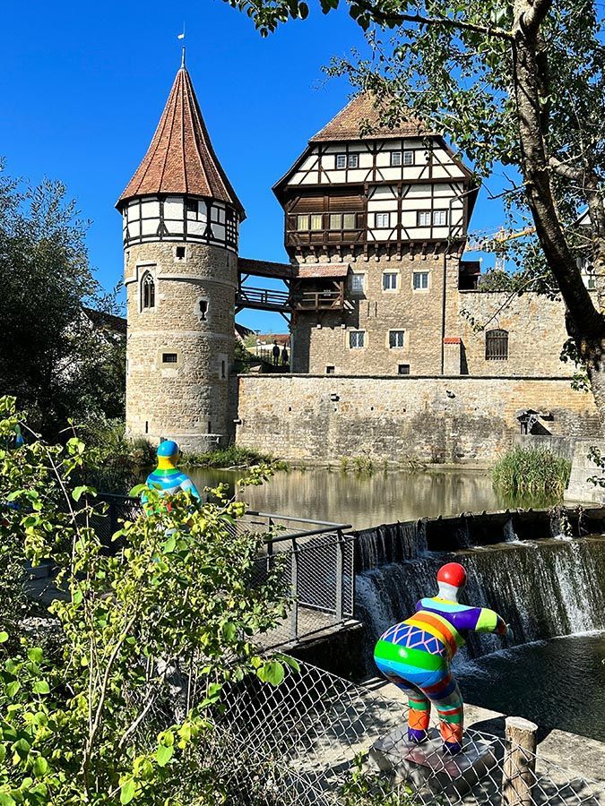 Zwei bunte Figuren in einem Burggraben neben einer mittelalterlichen Burg und einem Turm unter blauem Himmel