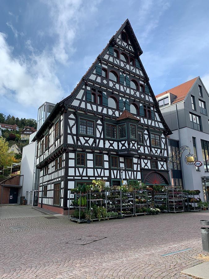 Fachwerkhaus mit Giebeldach, weißen Wänden und dunklen Balken auf einem gepflasterten Platz unter blauem Himmel