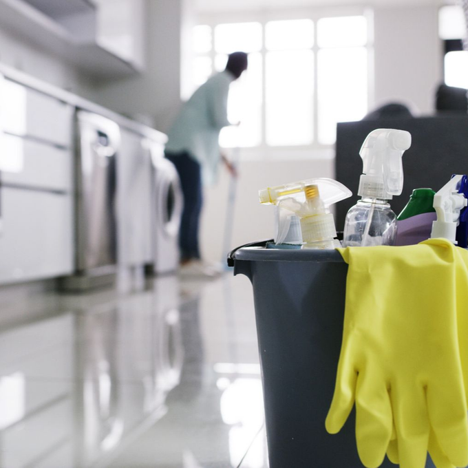 Des produits de nettoyage dans un seau, des gants jaunes dans une cuisine lumineuse, une personne qui nettoie en arrière-plan