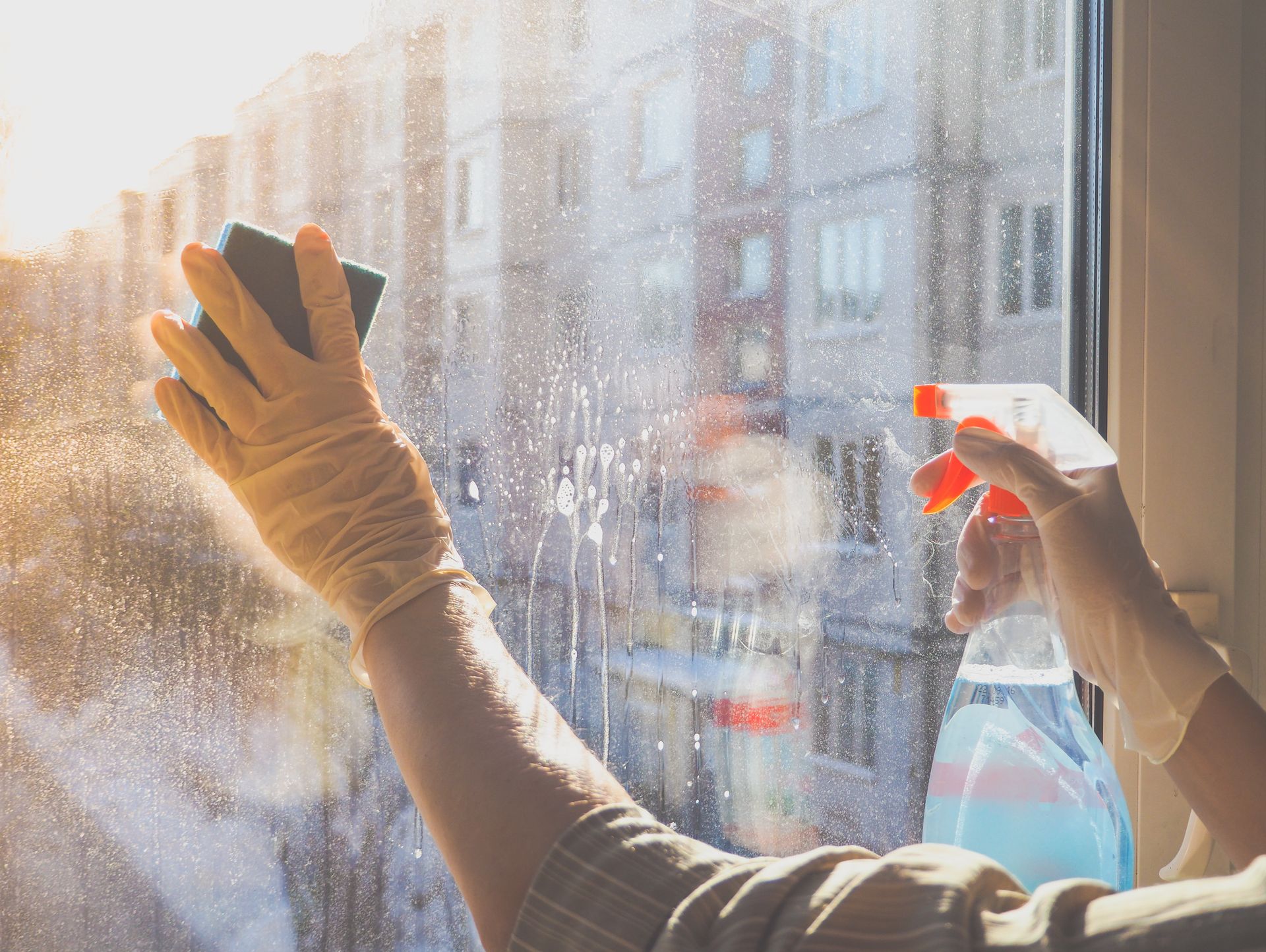 Des mains gantées nettoient une vitre ensoleillée avec un vaporisateur et un chiffon ; les immeubles de la ville se floutent à l’extérieur.