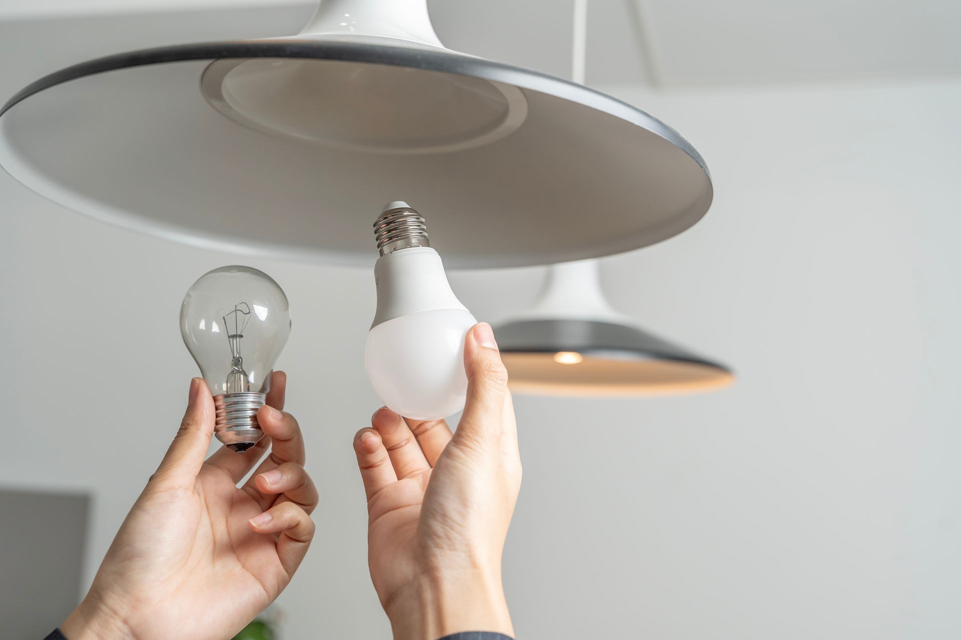 Des mains comparent une vieille ampoule transparente et une ampoule LED blanche sous un plafonnier.