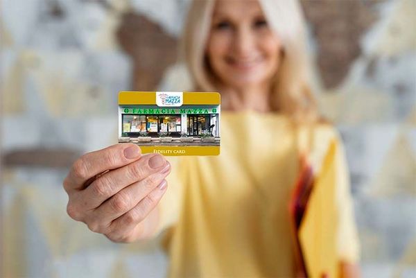 Farmacista in camice bianco sorride, braccia incrociate, in una farmacia con scaffali pieni di medicinali.