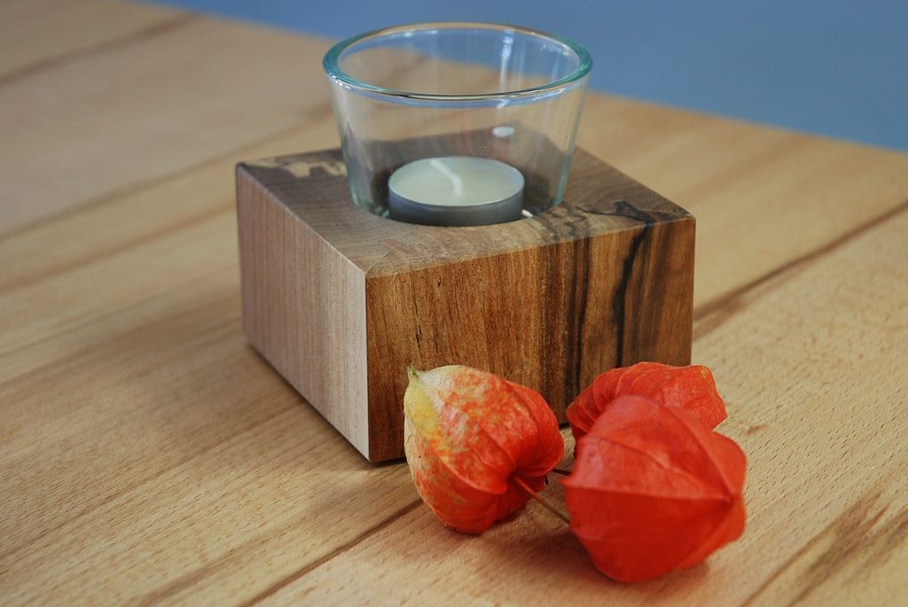 Ein hölzerner Kerzenhalter mit einer Kerze darin steht auf einem Holztisch neben zwei roten Blumen.