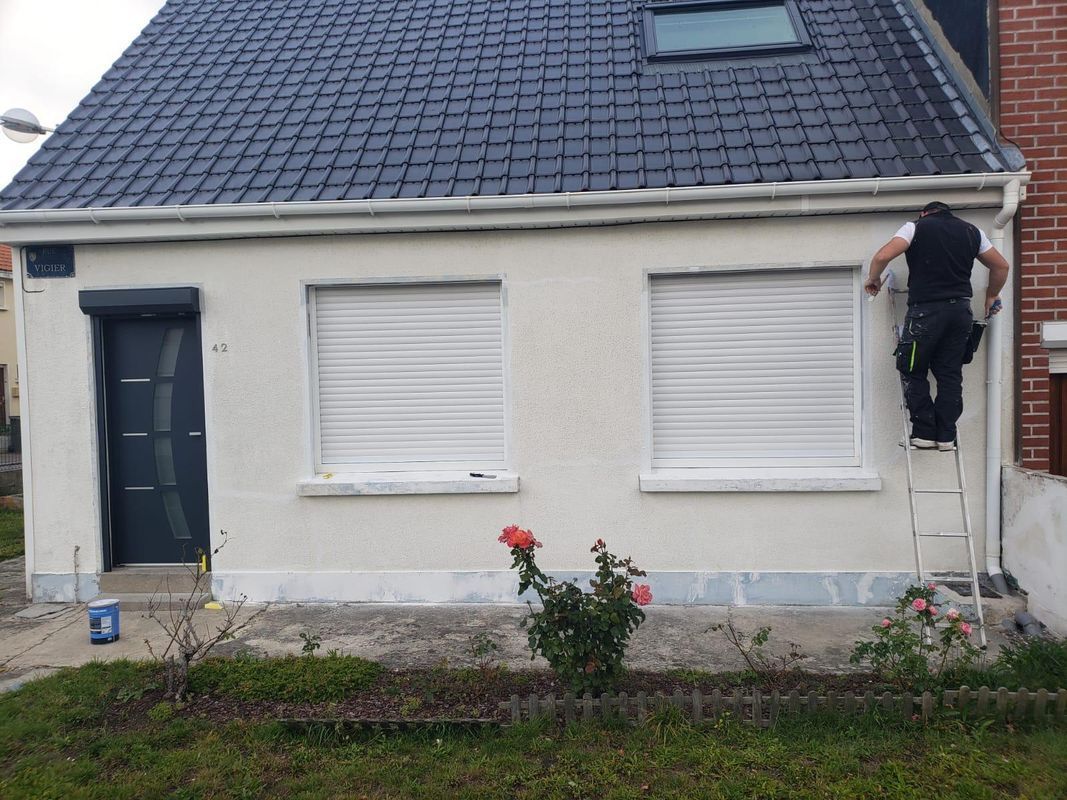 Personne sur une échelle nettoyant la gouttière d'une petite maison aux murs blancs, aux volets et au toit sombre.