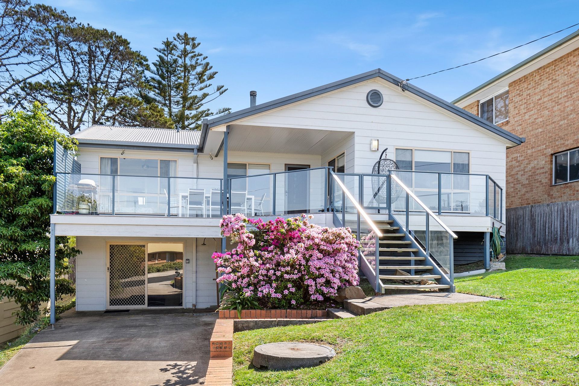 White house with glass balcony, pink flowers, and grassy lawn on a sunny day.
