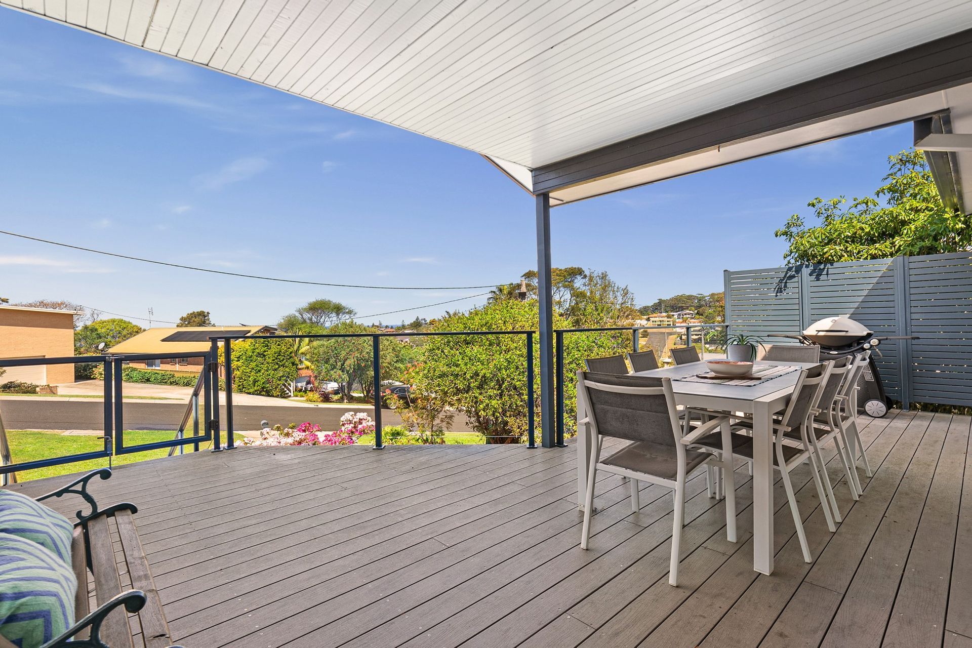 Deck with dining table, grill, and view of houses and greenery under a sunny, blue sky.