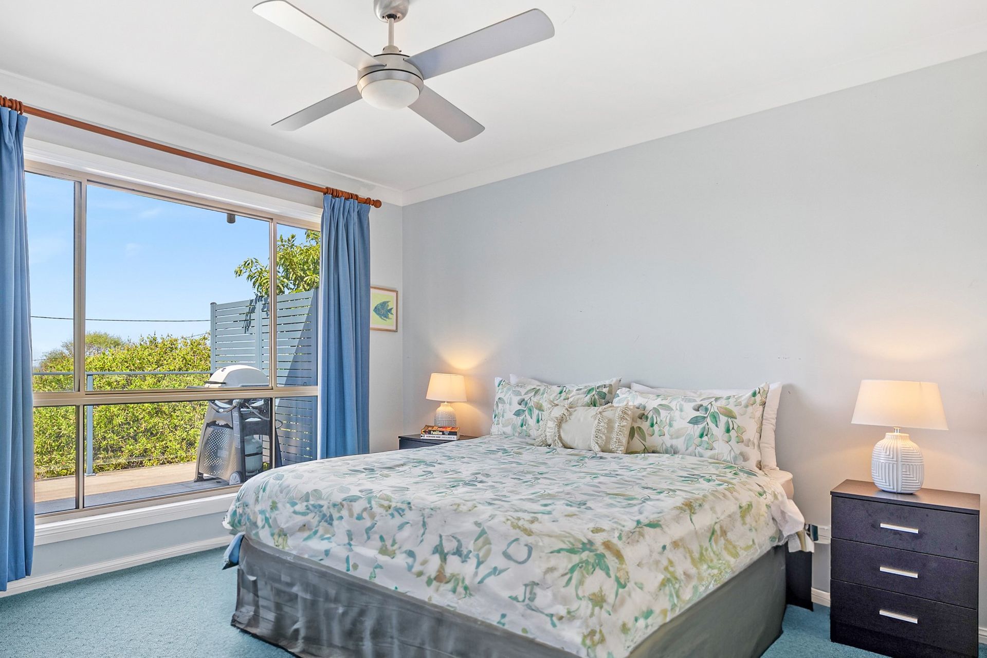 Bedroom with bed, blue curtains, large window, two bedside tables with lamps, light blue walls, and a ceiling fan.