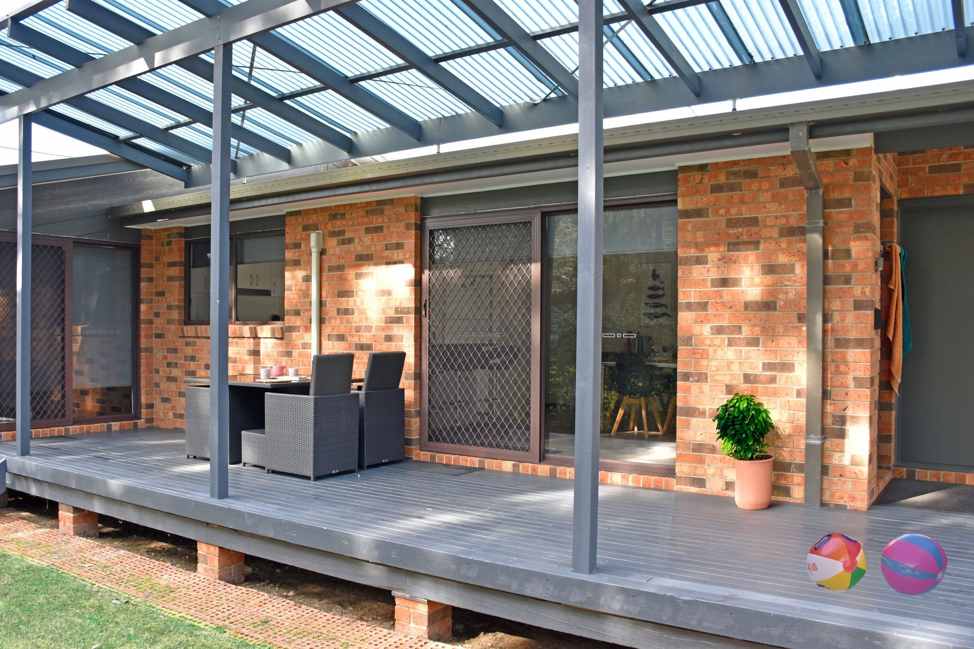 Patio with a brick building, covered by a corrugated roof. Dark gray decking, outdoor furniture, potted plant.