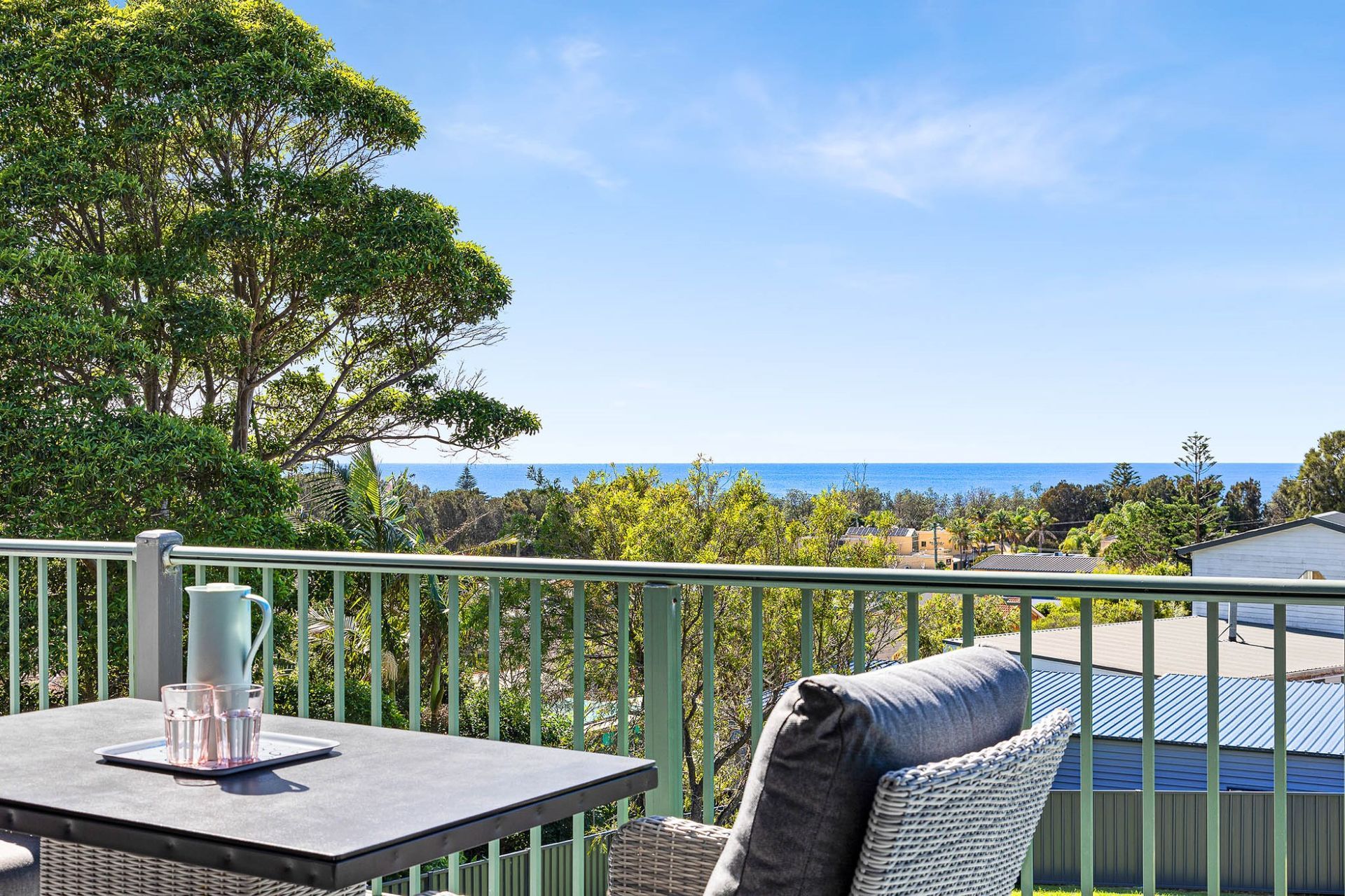 Balcony with table and chair overlooking trees, rooftops, and the ocean under a blue sky