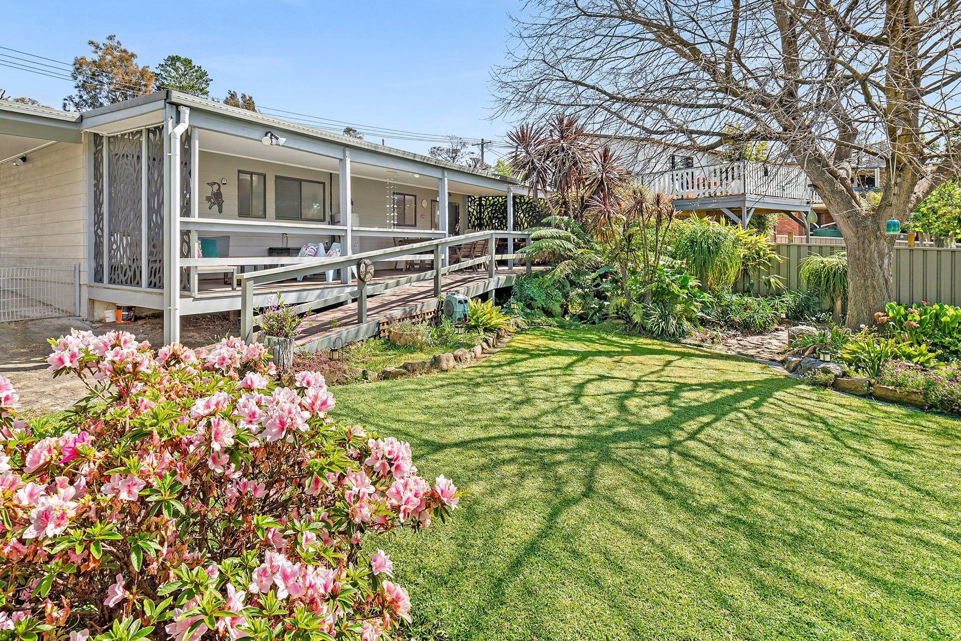 Sunlit backyard with green lawn, pink flowers, and a covered porch beside a house