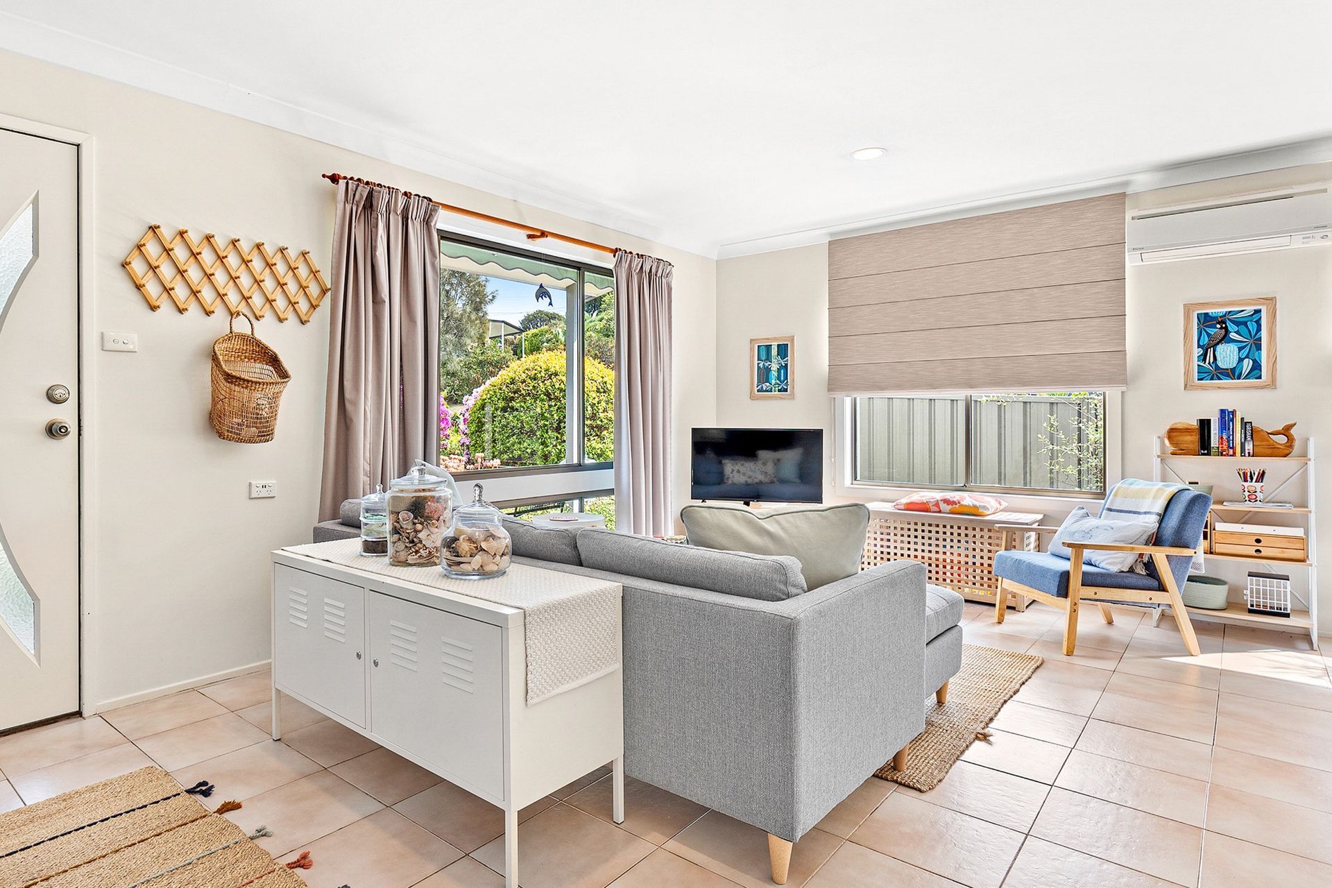 Bright living room with gray sofa, white chair, large window, and wooden dining table in a sunlit home.