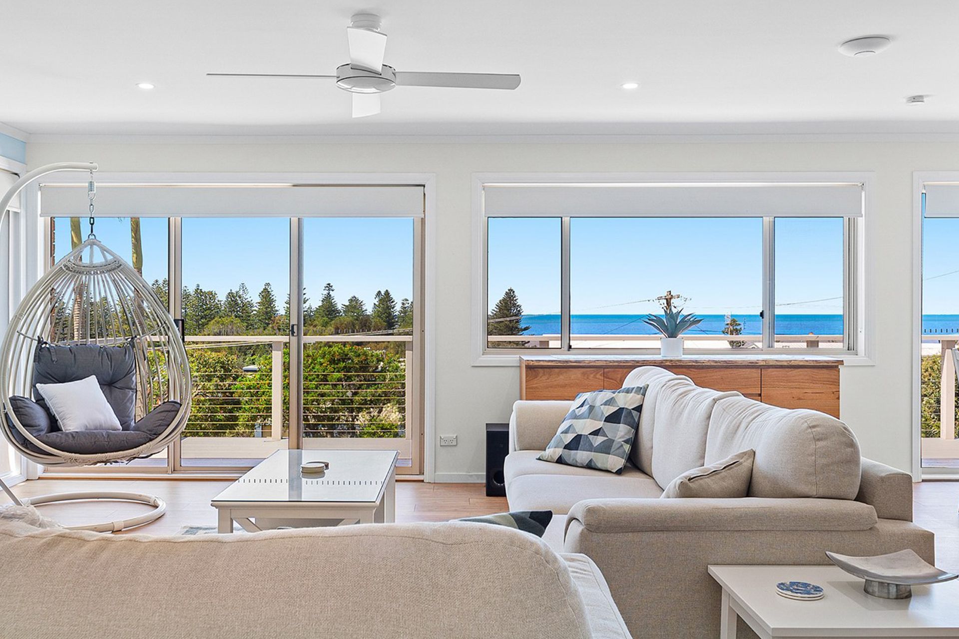 Bright coastal living room with white sofa, hanging chair, and ocean view through large windows