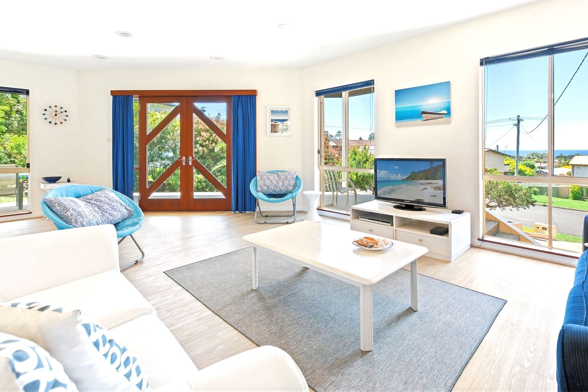 Bright living room with white furniture, blue accents, TV, coffee table, and ocean-view windows