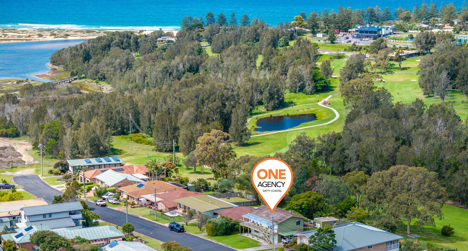 Aerial view of a coastal golf resort with ocean, green fairways, homes, and a One Agency sign