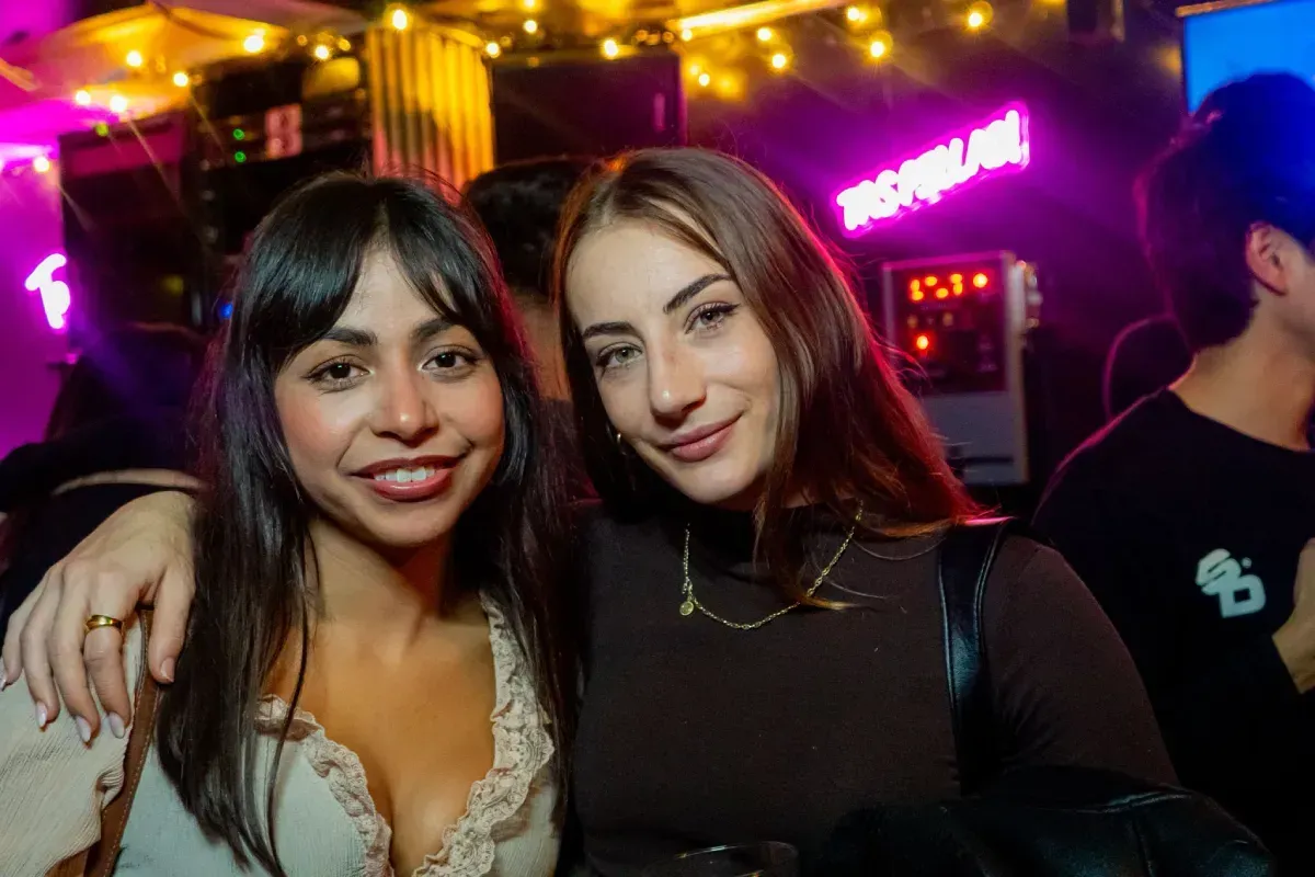 Dos mujeres sonriendo juntas en un bar poco iluminado, con luces de neón de fondo.