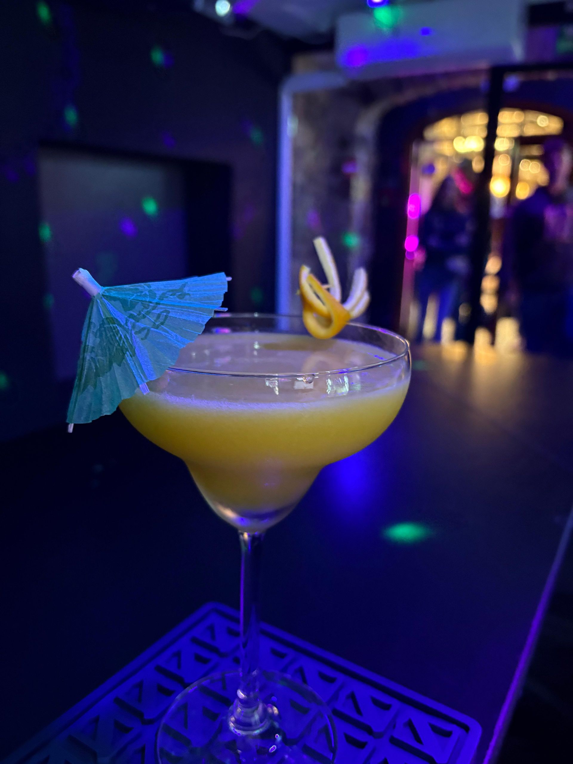 Cóctel en una copa con tallo y sombrilla azul sobre una barra de bar tenuemente iluminada, con luces de colores al fondo.