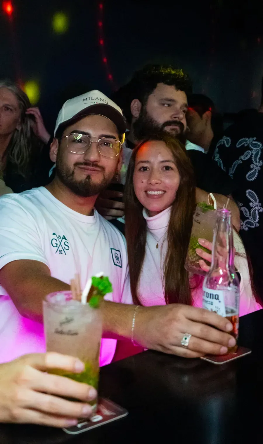 Pareja en un bar, tomando cócteles. El hombre lleva sombrero y gafas, la mujer sonríe. Ambiente oscuro con otras personas.