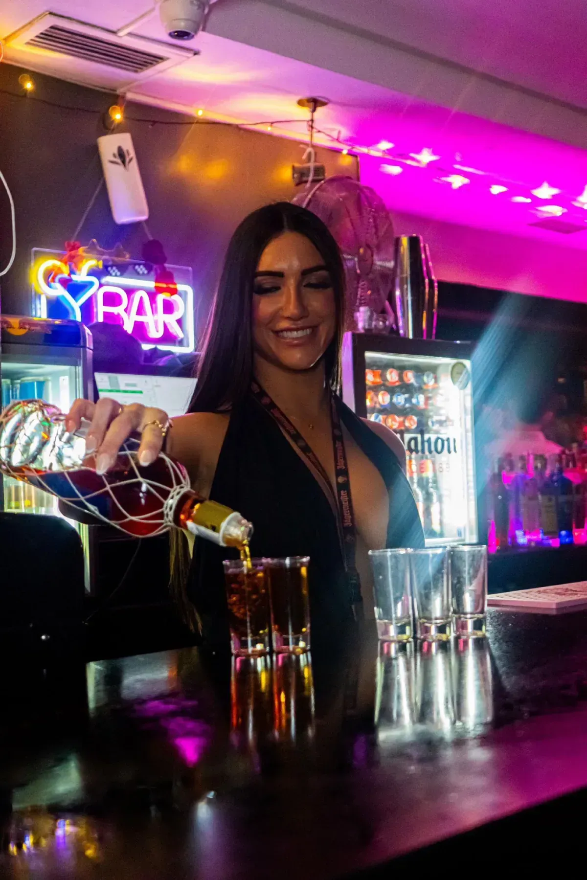 Camarero sirviendo licor en vasos de chupito en un bar, iluminado por luces de neón.