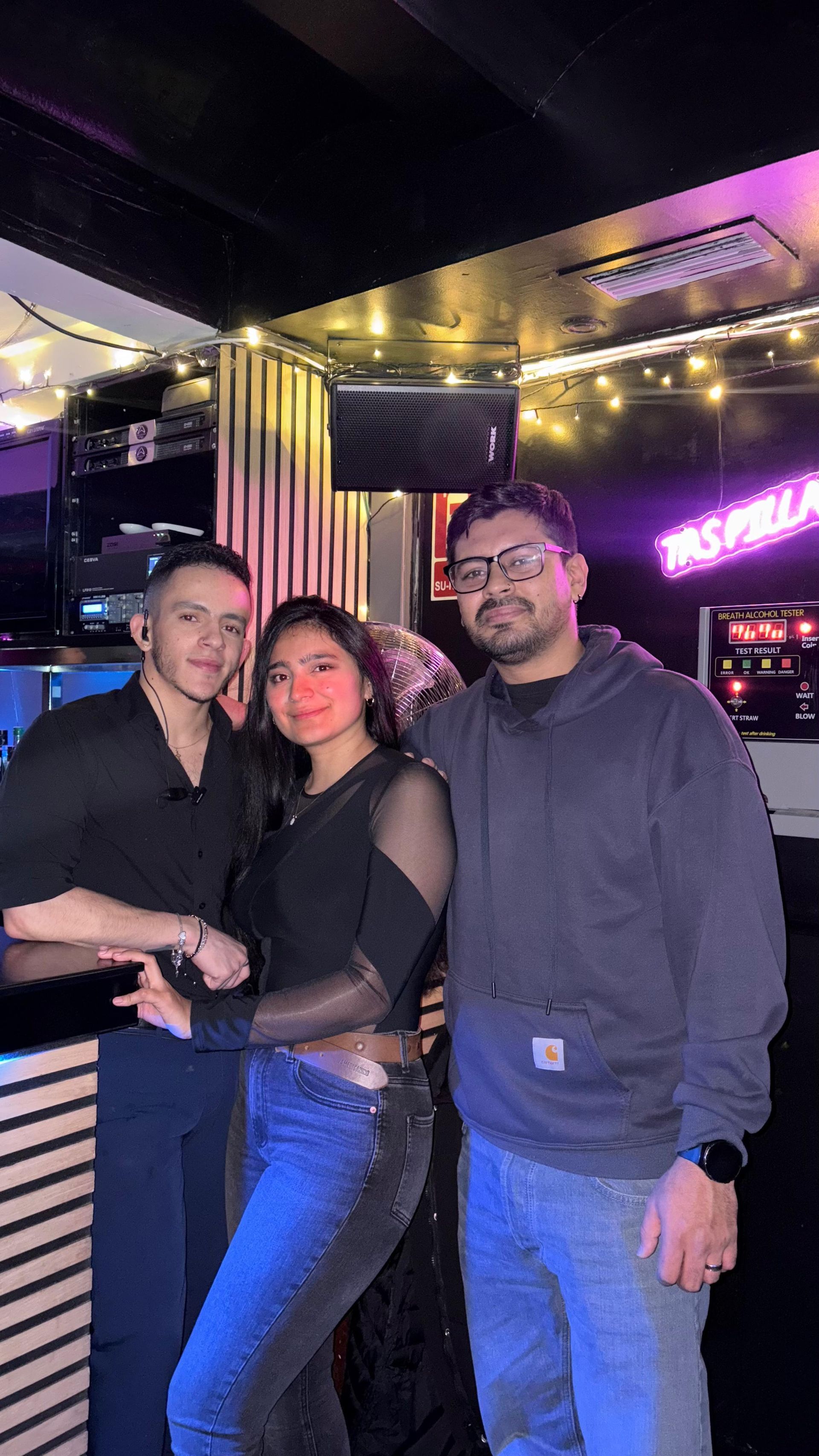 Tres personas posando juntas en un bar con poca luz, sonriendo bajo luces de neón moradas.