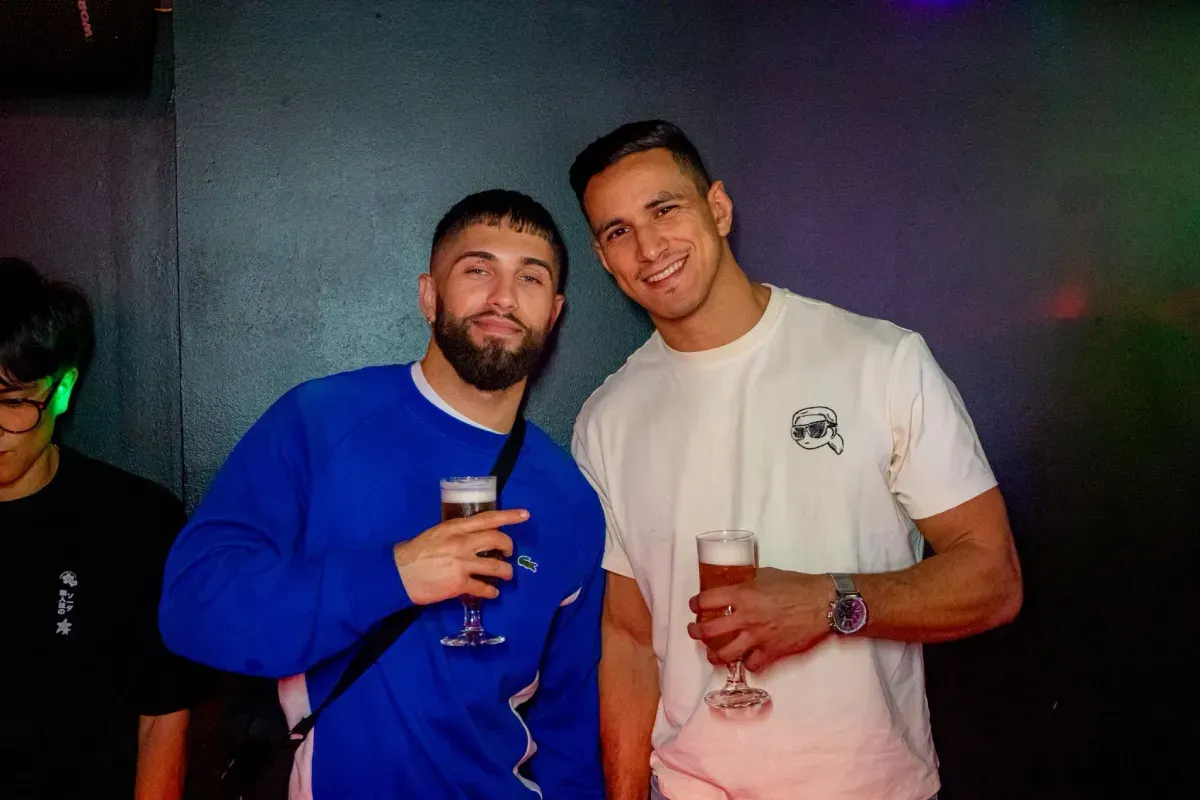 Dos hombres sonriendo, sosteniendo bebidas, dentro de un lugar oscuro con iluminación colorida.