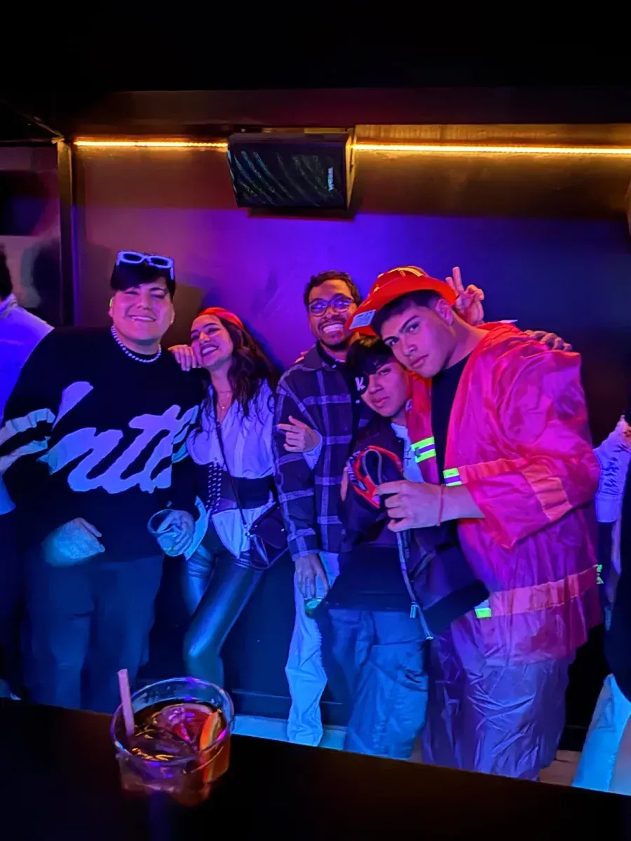 Grupo de personas en un bar, algunas con bebidas. Iluminación de neón e interior oscuro.