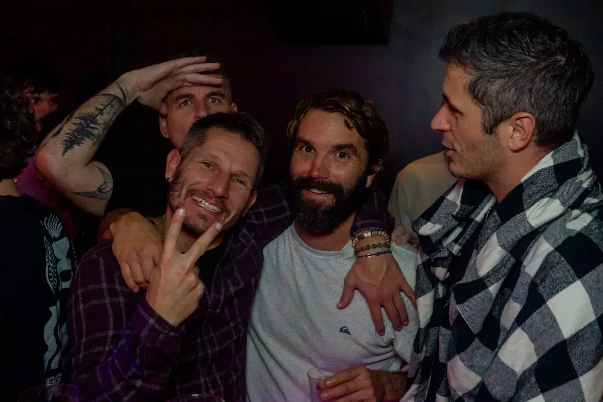 Cuatro hombres sonriendo en una fiesta, uno haciendo el signo de la paz, otros con los brazos alrededor del otro