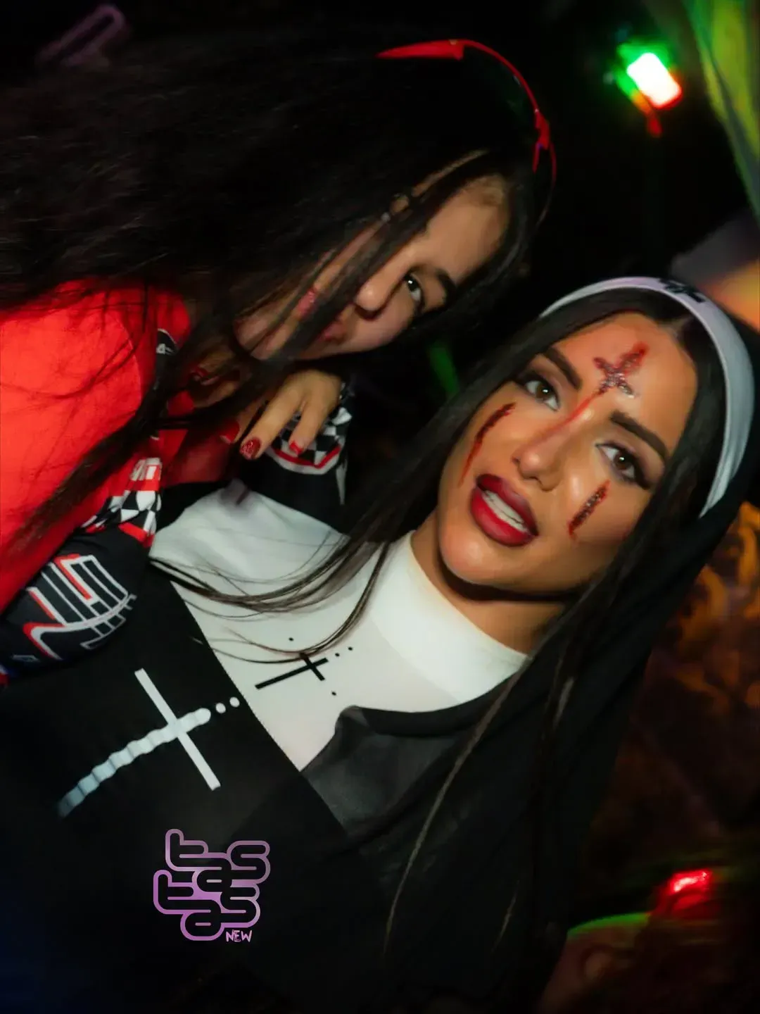 Dos mujeres disfrazadas de Halloween en una fiesta; una como monja con una cruz ensangrentada, la otra con un traje rojo.