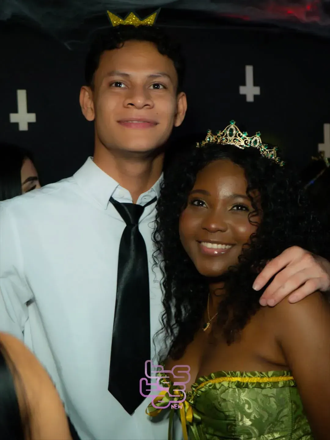 Dos personas disfrazadas sonríen a la cámara; el hombre lleva corbata, la mujer una corona, con un fondo negro.