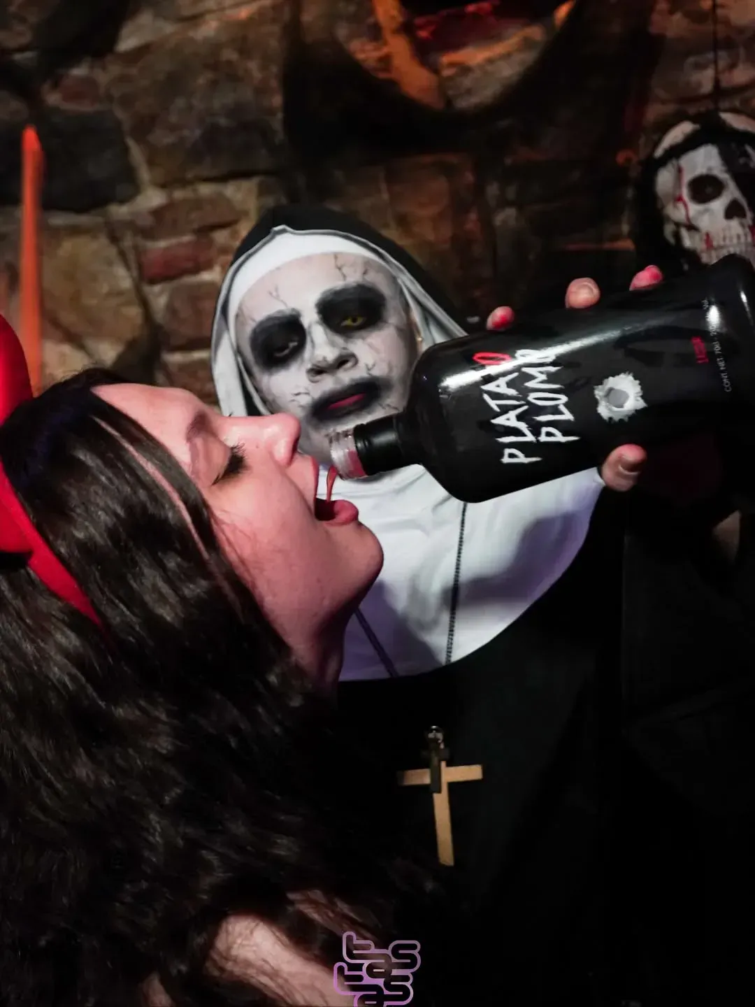 Persona vestida de monja vertiendo licor en la boca de una persona vestida de diablo, ambos en un entorno poco iluminado.