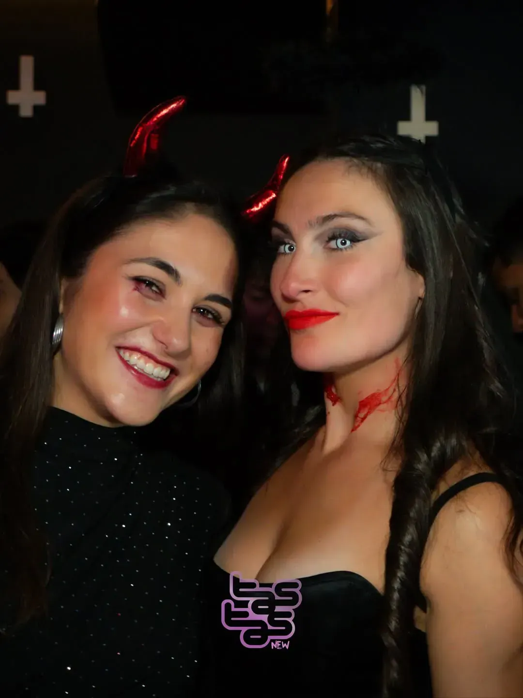Dos mujeres con disfraces de diablo, cuernos rojos y maquillaje sangriento en un evento oscuro.