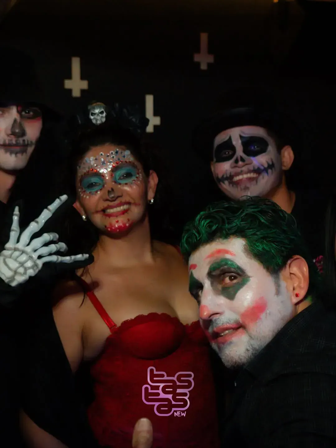 Grupo de personas disfrazadas en una fiesta: maquillaje de esqueleto, Joker y vestido rojo. Fondo negro con símbolos.