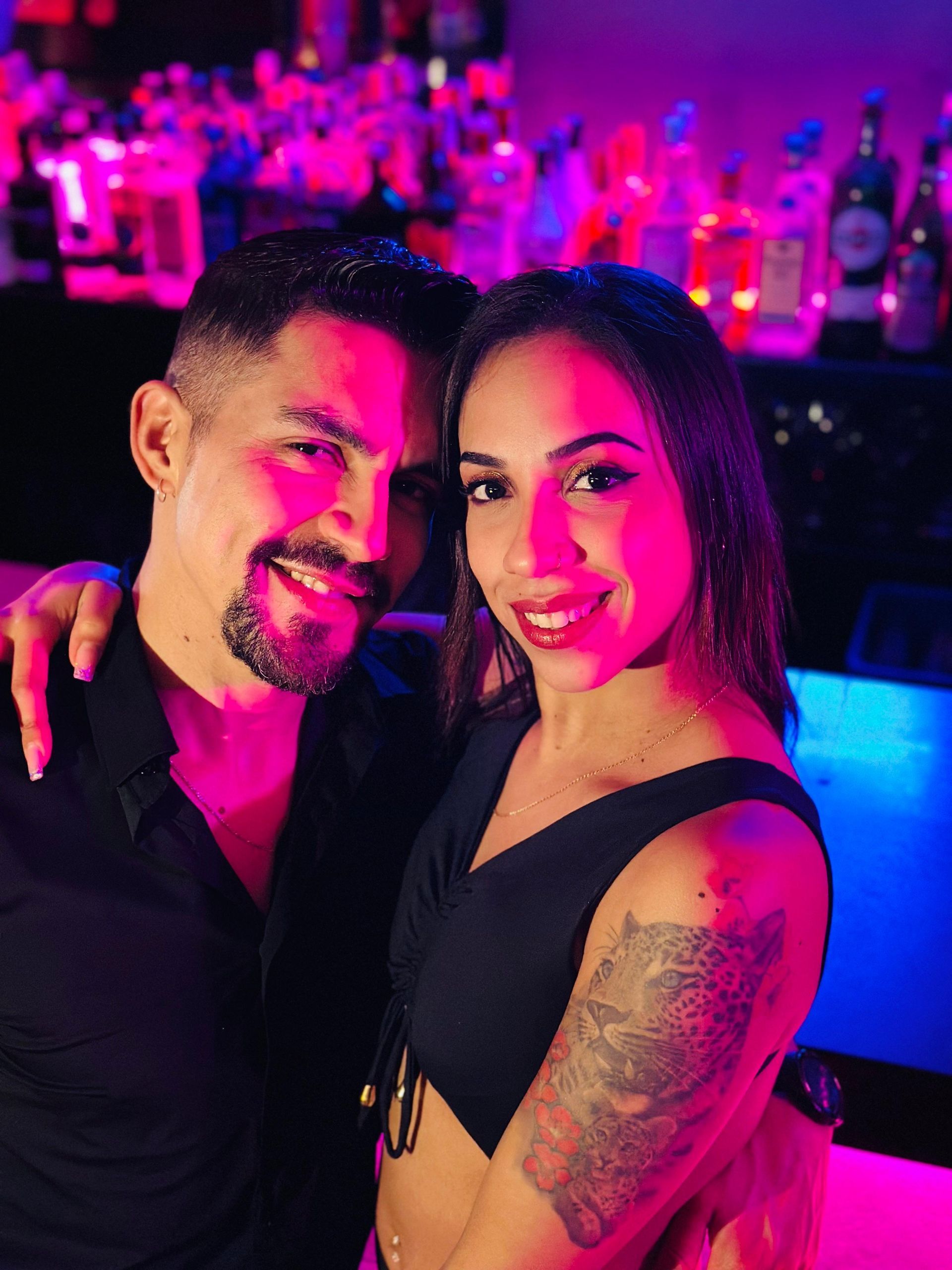 Dos personas posando juntas en un bar iluminado con luces de neón, sonriendo y abrazadas.