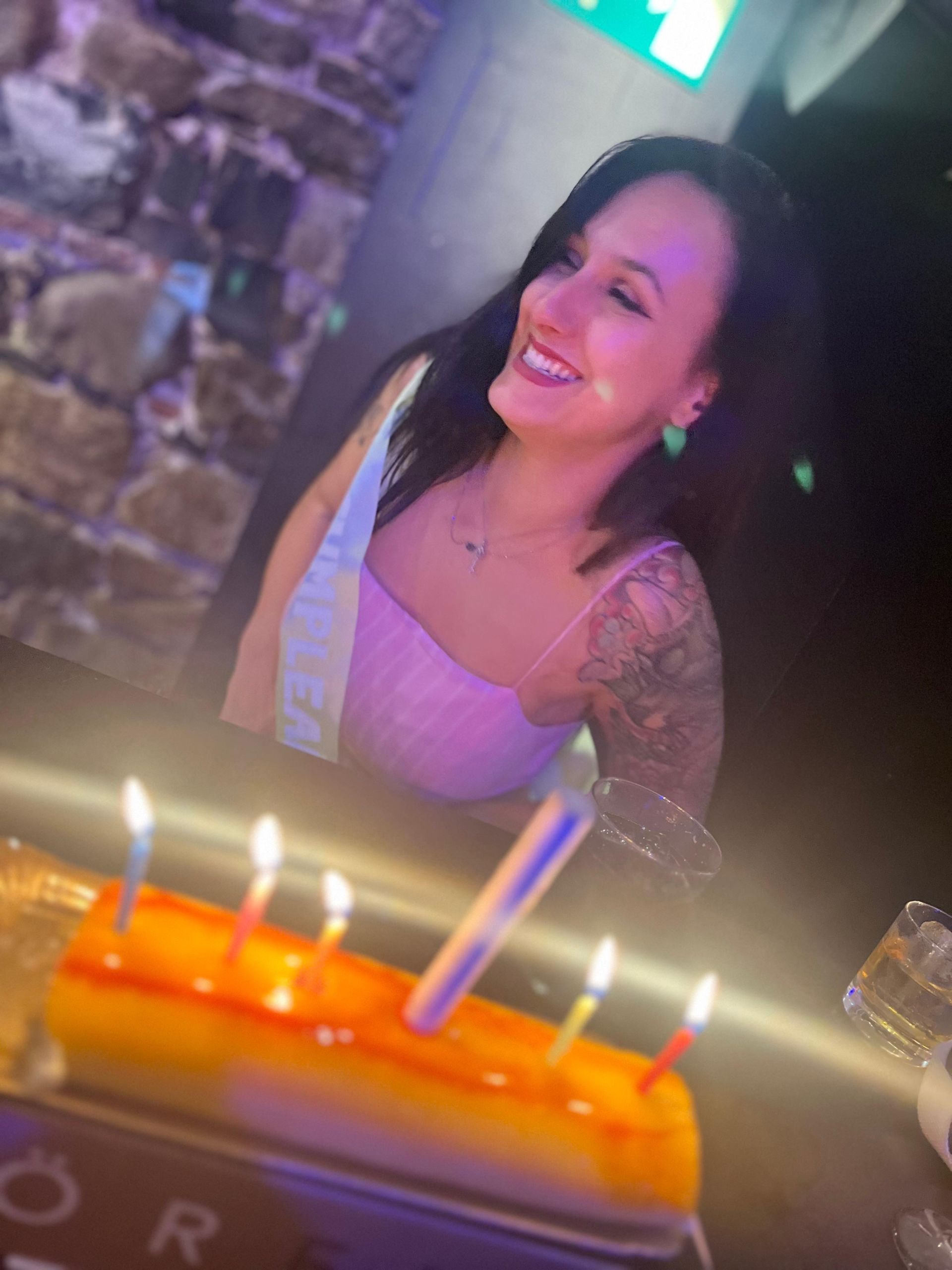 Mujer sonriente en una mesa de cumpleaños con velas encendidas, vestida con una blusa rosa claro y una banda.